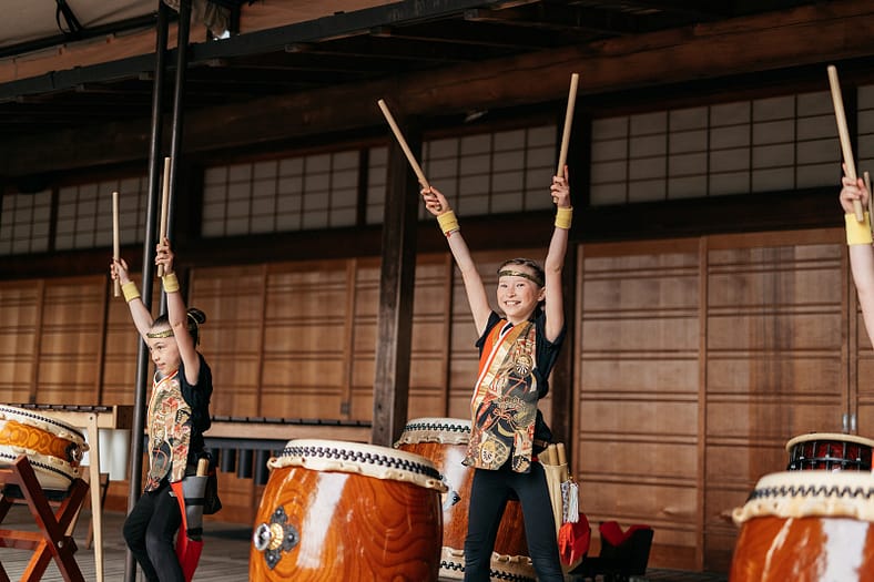 A child performer playing taiko drum smiles as she acknowledges an unseen audience.